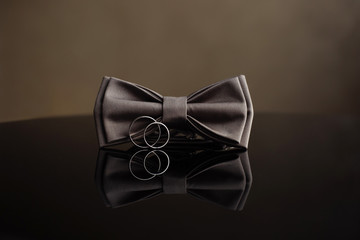 Wedding rings with a grooms bowtie on a mirrored table.