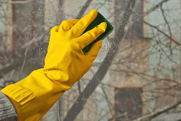 Hand in yellow rubber glove close up. The cleaner washes the window with a sponge. City street and buildings outside the window. The architecture is blurred in the background.