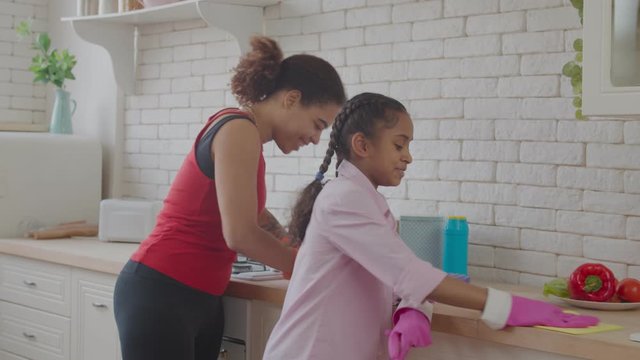 Industrious attractive african american mother and smiling adorable preadolescent mixed race daughter in rubber gloves cleaning countertop with detergent in domestic kitchen during spring cleanup.