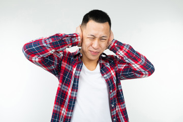 handsome asian guy in a checkered shirt wide open covers his ears from noise on a white studio background