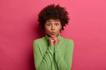 Fototapeta na wymiar Good looking young ethnic woman keeps lips rounded, keeps hands under chin, looks directly at camera, has widely opened eyes, dressed in casual wear, poses against pink background, waits for kiss