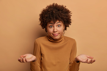 Puzzled unaware woman with Afro hairstyle shrugs shoulders, spreads palms and looks doubtfully, has...