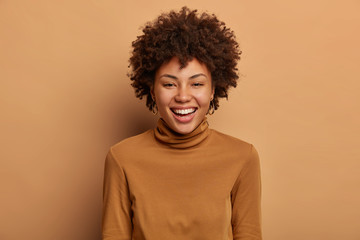 Obraz na płótnie Canvas Pretty merry woman with Afro hair, healthy skin, white beaming smile, enjoys hearing funny stories, expresses positive emotions and feelings, wears casual outfit, models indoor. Happiness concept