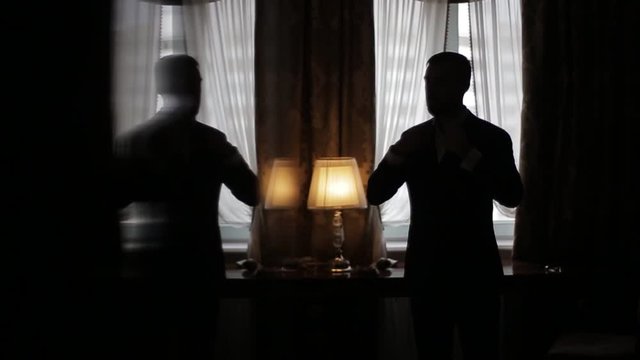 Silhouette Of Stylish Wealthy Man Standing In Dark Room By The Window And Turned On A Beautiful Lamp And Straightening His Expensive Suit In An Expensive Hotel Room. Preparing For A Business Meeting.