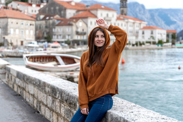Fototapeta na wymiar Woman in orange hoodie stands by the sea in city Perast on the background in Montenegro