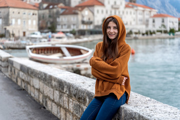 Fototapeta premium Woman in orange hoodie stands by the sea in city Perast on the background in Montenegro