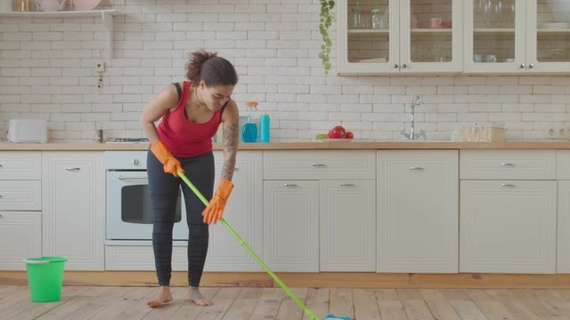 Positive industrious attractive african american woman in rubber gloves with bucket mopping kitchen floor during cleanup. Housekeeping mixed race female washing floor with mop and cleaning detergent.