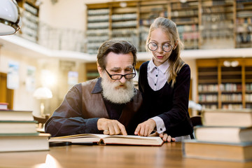 Happy international literacy and book day and knowledge concept. Handsome senior bearded grandfather reading book together with his cute pretty granddaughter, pointing on interesting moment in book