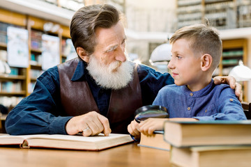 Happy smiling little boy with his cheerful bearded grandfather reading books at library, looking...