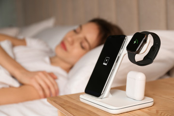 Smartphone, watch, earphones charging on wireless pad and sleeping woman in room