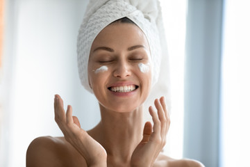 Close up head shot beautiful lady laughing while applying skincare moisturizing creme after morning showering. Happy young woman with wrapped in towel head enjoying smoothing skin, domestic routine. - obrazy, fototapety, plakaty