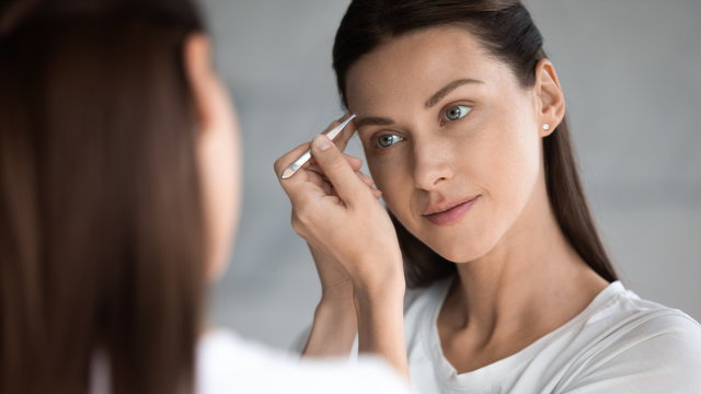 Mirror reflection attractive young brunette woman using tweezers, doing correction procedure in bathroom, satisfied with long lasting styling or perfect eyebrows shape, face treatment concept.