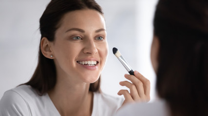 Close up head shot beautiful young brunette woman looking in mirror, using brush, doing make up in morning. Smiling pretty lady satisfied with skin condition, applying foundation in bathroom.