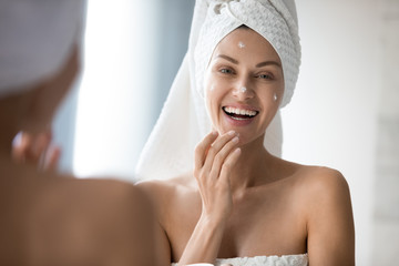 Attractive young lady with towel on head applying moisturizing smoothing cream on face after...