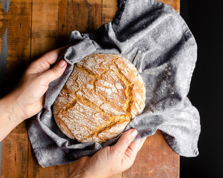 Hands Placing A Freshly Baked Loaf Of Bread Wrapped In A Grey Cloth Onto A Wood Tabletop.