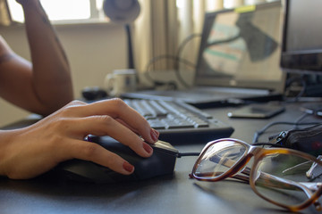 Closeup on the hand of a woman using her computer and glasses, with a keyboard, laptop and monitor...