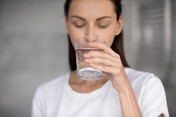 Focus on peaceful young brunette woman drinking glass of fresh aqua, head shot close up. Attractive 30s lady sipping water, enjoying daily healthcare routine, every day healthy habit concept.