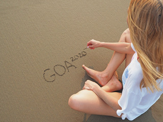 A girl sits on the sand facing the sea and writes the inscription goa 2020 in the sunset rays - obrazy, fototapety, plakaty