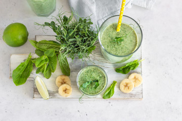 Green smoothie with spinach, banana, lime, mint and young sprout microgreen on light grey stone background.