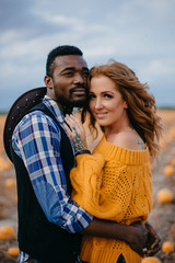 An African man and a Caucasian woman are standing embracing in a pumpkin field and looking at the camera. Close-up portrait.