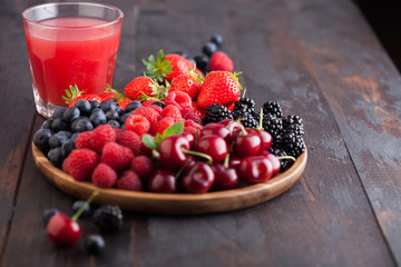 Fresh organic summer berries mix in round wooden tray with glass of juice on dark wooden table background. Raspberries, strawberries, blueberries, blackberries and cherries. Space for text