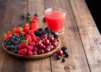 Fresh organic summer berries mix in round wooden tray with glass of juice on light wooden table background. Raspberries, strawberries, blueberries, blackberries and cherries. Space for text