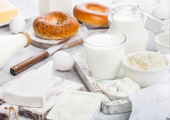 Fototapeta na wymiar Fresh dairy products in vintage wooden box on white table background. Jar and glass of milk, bowl of sour cream and cheese and eggs. Fresh baked bagel on round chopping board with knife.