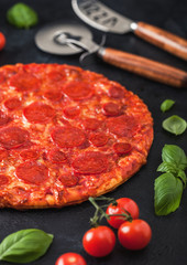 Fresh round baked Hot and Spicy Pepperoni pizza with wheel cutter and knife with tomatoes and basil on black background with kitchen cloth. Close up