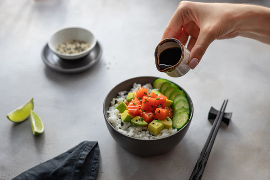 Woman's Hands Holding A Saucepan With Soy Sauce To Sprinkle A Hawaiian Poke Bowl With Salmon, Rice, Avocado And Cucumber. Traditional Asian Food. Horizontal Image, Gray Background.