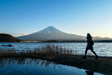 A woman walking at the side of Kawaguchiko Lake and watching Mt Fuji, Japan. Reflection of the woman in the water. Top of volcano covered with snow. Exploring new places. Serenity. Soft sunset colors