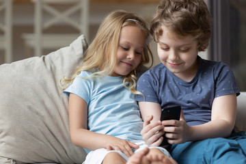 Little clever brother and sister sit on couch using cellphone browsing web together, happy small children relax on comfortable sofa at home watch video or cartoon on smartphone, technology concept