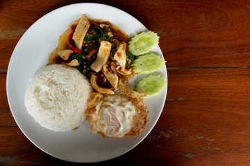 Top view of Rice topped with stir-fried octopus and basil and egg. On a white plate resting on a wooden table.