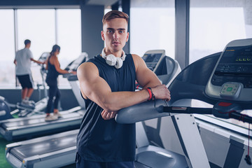 Smiling positive confident male personal instructor with arms crossed arms near treadmill at gym in fitness gym.