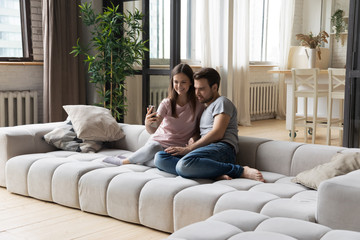 Obraz premium Happy young couple sit on big comfortable couch in new flat making self-portrait picture on modern gadget cellphone, smiling husband and wife rest on sofa take selfie on smartphone together