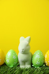 Easter bunny toy and dyed eggs on green grass against yellow background. Space for text