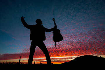 Silhouette of male musician standing, holding guitar on a scenic violet sunset sky 