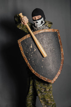 A radical protesting man in camouflage uniform with a balaclava in the form of a skull on his head holds a shield and a baseball bat in his hands. Calls for battle. Studio photo on a gray background.