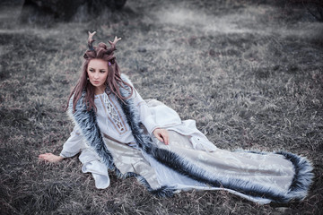 Portrait of amazing cute young woman in fairy tale image with long hair in interesting hairstyle and horns on her head in beige dress with gray fur sitts on grass