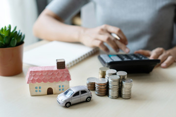Close up hand using calculator, stack of coins, toy house and car on table, saving for future,...