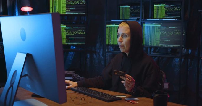 Caucasian female hacker in hood sitting at desk in front of big computer screen and holding credit card. Woman buying online and typing card number as paying. At night in monitoring room.