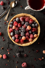 Healthy pastry concept. Delicious open pie with frozen raspberries, blackberries and black currants served with hibiscus tea and dry flowers on black background