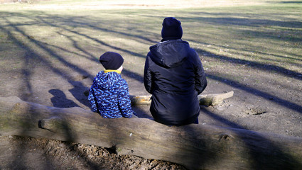 Fototapeta Mama z synem na pniu drzewa odpoczywa po jesiennym spacerze. Ubrani w ciepłe kurtki i czapki, w czasie podróży, spaceru po lesie, wyprawy w góry. Wspólne spędzanie czasu z dzieckiem. obraz