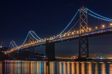 San Francisco Bay Bridge at night, lit up by yellow and blue lights, reflecting of the water in the Bay, long exposure