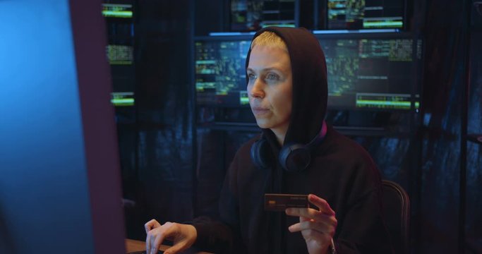 Joyful Caucasian woman in hood typing credit card number on keyboard when shopping in internet at big computer screen in dark IT monitoring room at night. Female hacker buying and paying online.