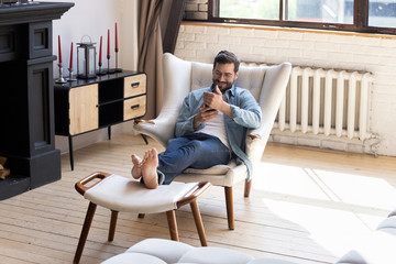 Smiling young man using smartphone, sitting in armchair in modern living room, happy male holding phone, looking at screen, chatting in social network or shopping online, playing game, using apps