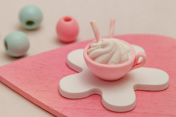 Products from polymer clay: a cup with a dessert with marshmallows, meringues and macaroons in pastel blue and pink colors on a decorative wooden heart.