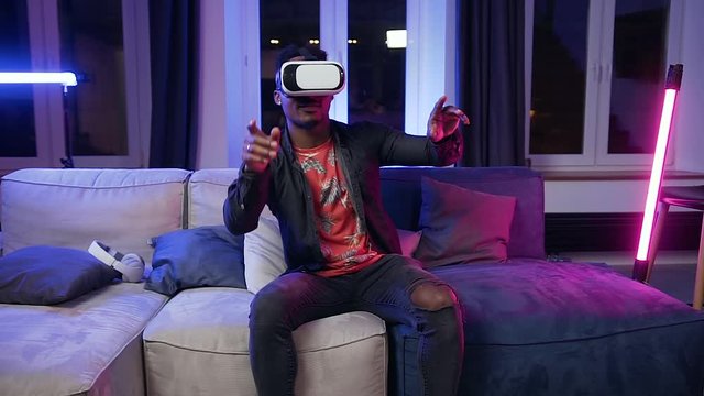 Attractive smiling young black-skinned man in virtual reality headset sitting on the couch in the contemporary room in the evening time