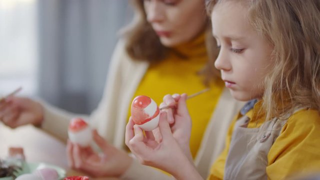 Side view of cute girl and young woman in yellow and beige outfits using brushes and red gouache to paint on white boiled eggs while preparing for Easter