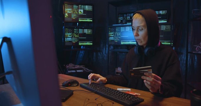 Caucasian female hacker in hood committing financial cyber crime while frauding with credit cards on computer in dark hacking room. Woman entering number and password wile typping on keyboard.
