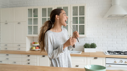Funny beautiful woman singing into beater, cooking in modern kitchen, holding whisk as microphone,...